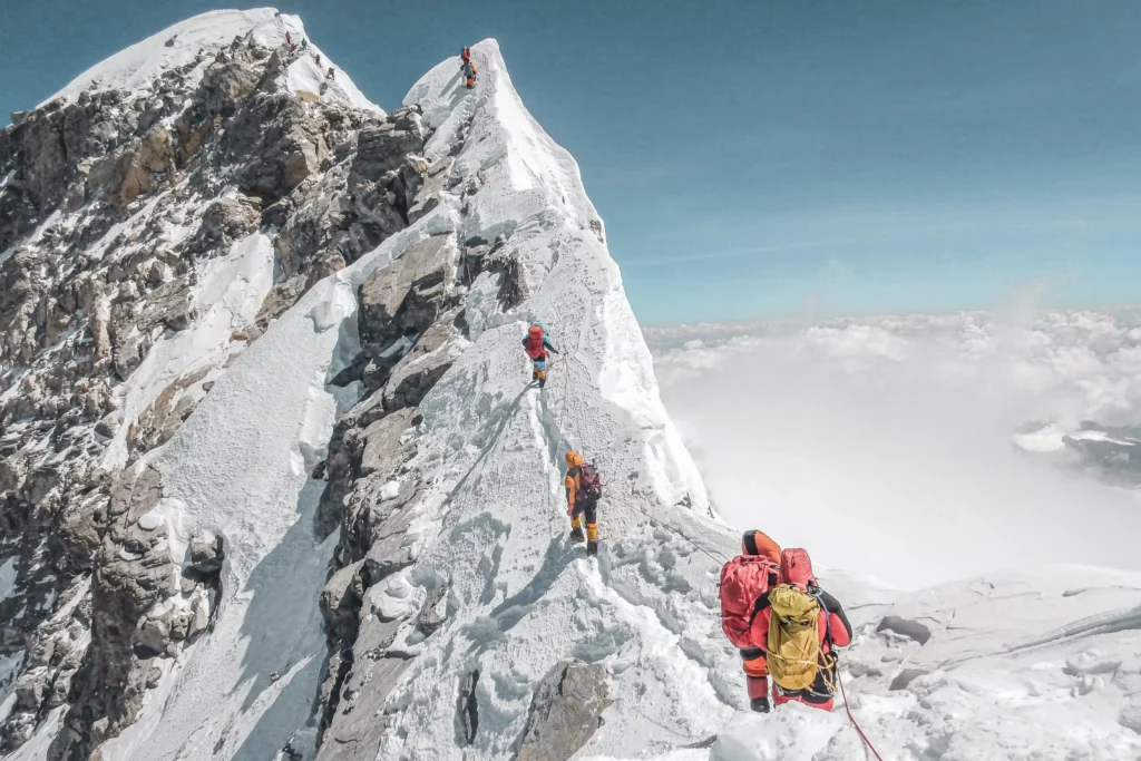 A group of climbers ascends a steep, snow-covered mountain ridge. They are roped together, wearing bright jackets and backpacks, with a clear blue sky above and clouds below, indicating a high altitude. The rocky, icy terrain is visible on both sides of the narrow path leading to the summit.