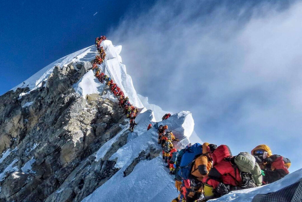 A long line of climbers in colorful gear ascends a steep, snow-covered mountain ridge under a clear blue sky. The climbers are roped together, navigating the icy and rocky terrain, with some resting or preparing near the bottom of the ridge. Clouds are visible below, indicating high altitude.