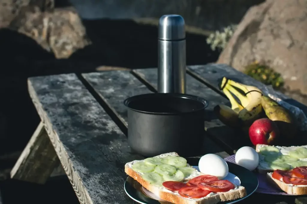 Outdoor camping breakfast setup on a rustic wooden picnic table with sandwiches topped with tomato and cucumber, boiled eggs, bananas, an apple, a thermos, and a cooking pot, surrounded by rocks in natural sunlight