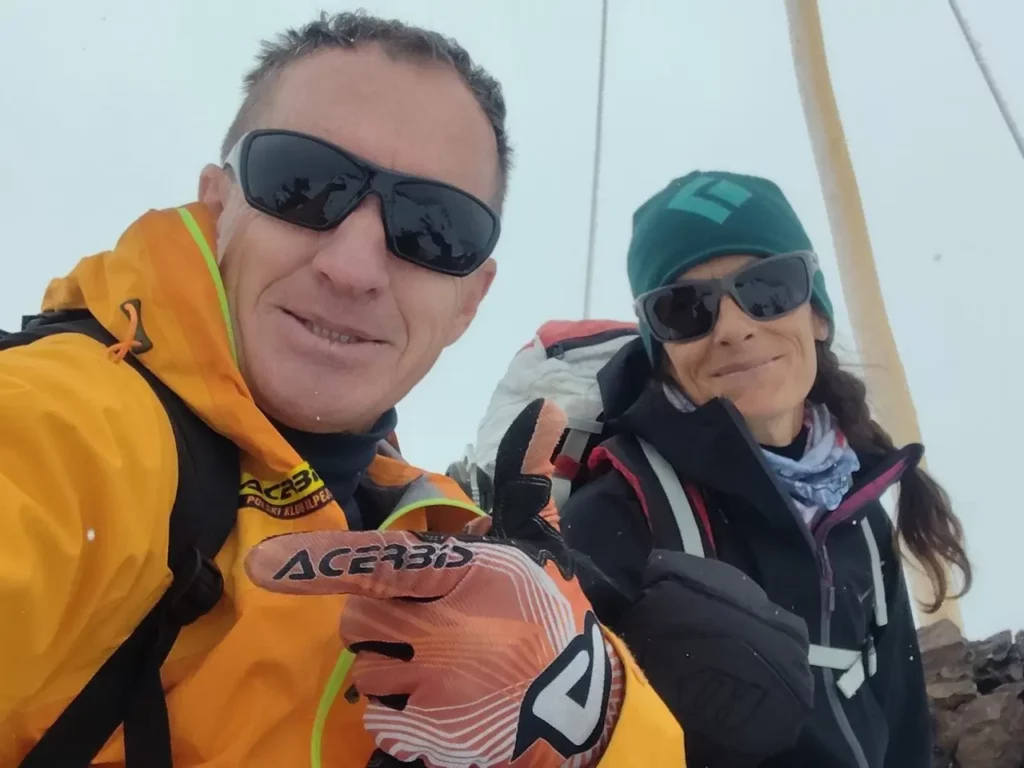 Two people wearing outdoor gear, including bright jackets, gloves, and sunglasses, pose for a selfie with a snowy mountain background. The person on the left is in a yellow jacket with an "Acerbis" glove raised, while the person on the right wears a green beanie and a dark jacket.