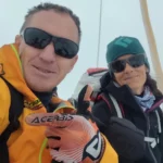 Two people wearing outdoor gear, including bright jackets, gloves, and sunglasses, pose for a selfie with a snowy mountain background. The person on the left is in a yellow jacket with an "Acerbis" glove raised, while the person on the right wears a green beanie and a dark jacket.