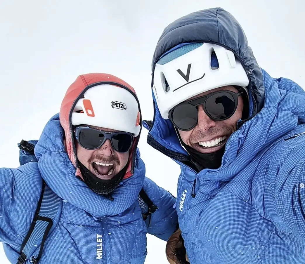 Two smiling climbers dressed in blue down suits and helmets take a selfie in snowy, high-altitude conditions. Both wear sunglasses and mountaineering gear, showing excitement and camaraderie during their expedition.