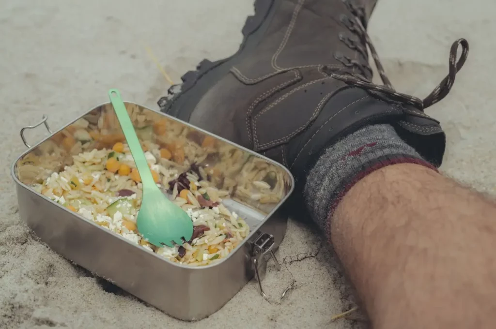 Close-up of a hiker’s meal in a metal container placed on sandy ground, featuring orzo pasta salad with chickpeas and vegetables, next to a hiking boot and sock-covered leg.