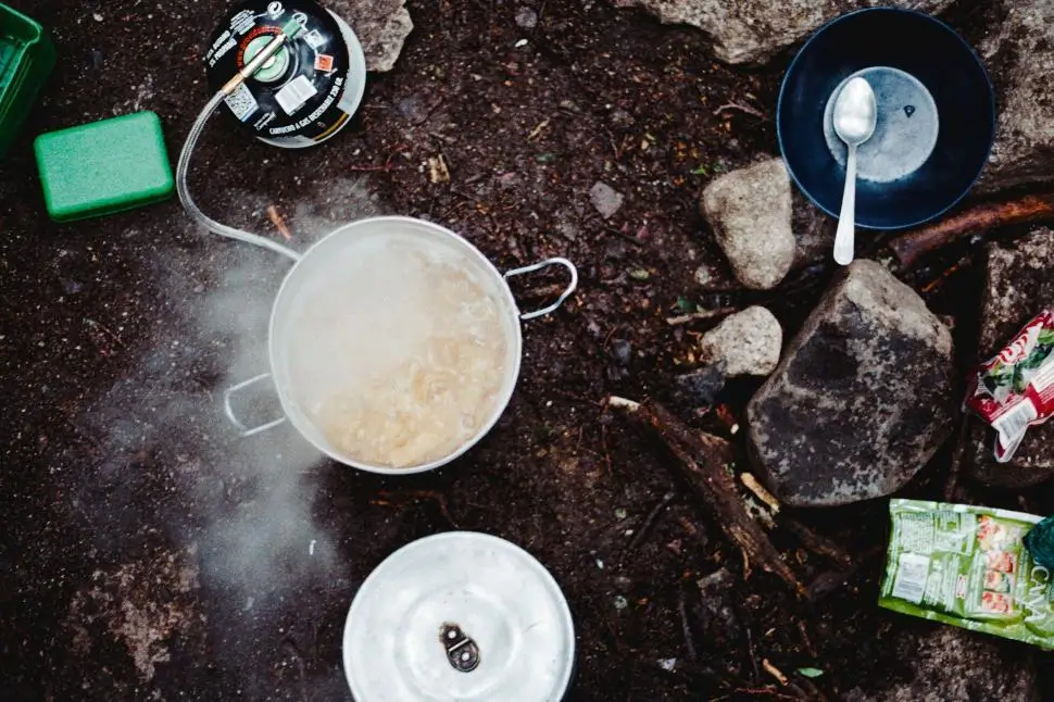 Overhead view of a camp cooking setup with a pot of boiling food on a portable gas stove, surrounded by rocks, a bowl with a spoon, food packaging, and outdoor gear on forest ground.