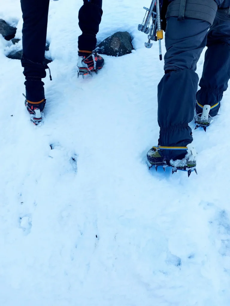 Close-up of two climbers' feet wearing crampons on a snowy surface, with climbing gear attached to their legs.