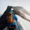 A skier with an orange helmet and backpack ascends a snow-covered slope, using ski poles, with rugged, snow-dusted mountains stretching into the distance under a cloudy sky.