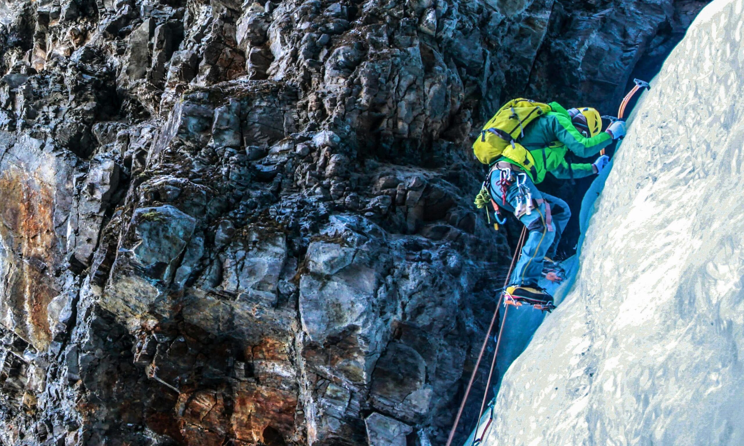 A climber wearing bright gear and a yellow helmet ascends a steep icy rock face using ice axes and crampons, secured by ropes against a rugged cliff.