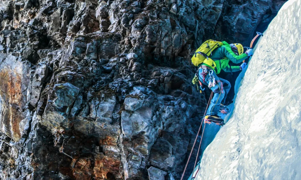 A climber in a green jacket and yellow helmet ascends a steep ice wall with an ice axe, secured by a rope, alongside a rugged rock face.