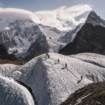 A detailed image of K2's southeast face showing the classic Abruzzi Spur route. The photo is marked with the climbing route in red, starting from K2 Base Camp at 16,500 ft and progressing through key points: Advanced Base Camp, Camp 1 (16,500 ft), Camp 2 (22,000 ft), Camp 3 (24,000 ft), Camp 4 (25,000 ft), and the summit at 28,251 ft. Landmarks such as the Black Pyramid, House’s Chimney, and Chinese Camp 1 (20,500 ft) are labeled along the ascent. The mountain is covered in snow and ice under a bright blue sky.