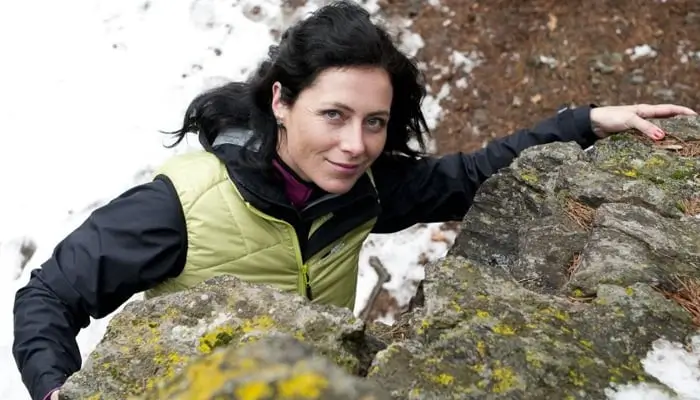 Klára Kolouchová climbing on rocky terrain with patches of snow, wearing a light green vest and black jacket, looking up and smiling at the camera.