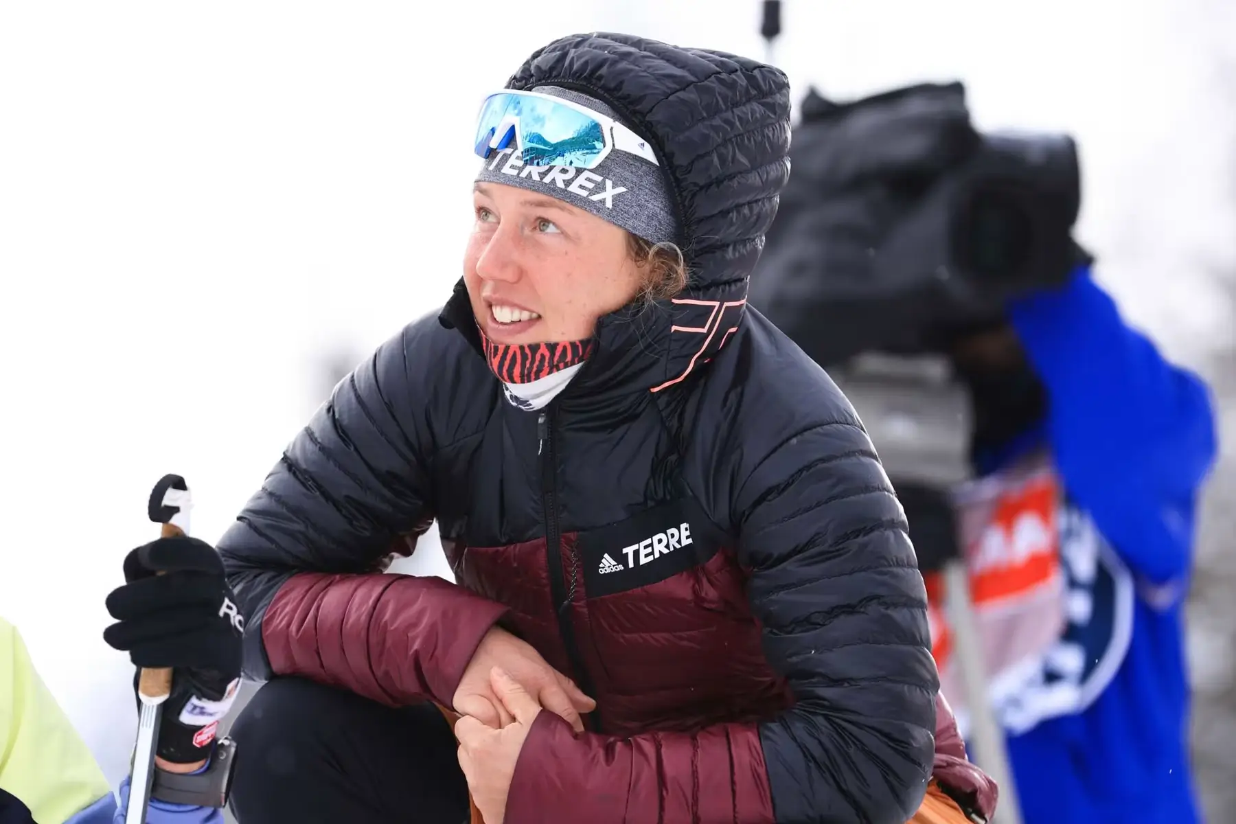 Laura Dahlmeier, dressed in a black and maroon insulated jacket with a TERREX logo, crouches in the snow smiling, wearing reflective ski goggles and a hood, during a winter sports event. A cameraman is visible in the blurred background.