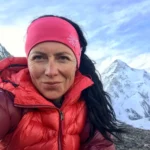 Klára Kolouchová, wearing a red down jacket and pink headband, smiles slightly while standing in a rocky alpine area with a snow-covered mountain in the background.