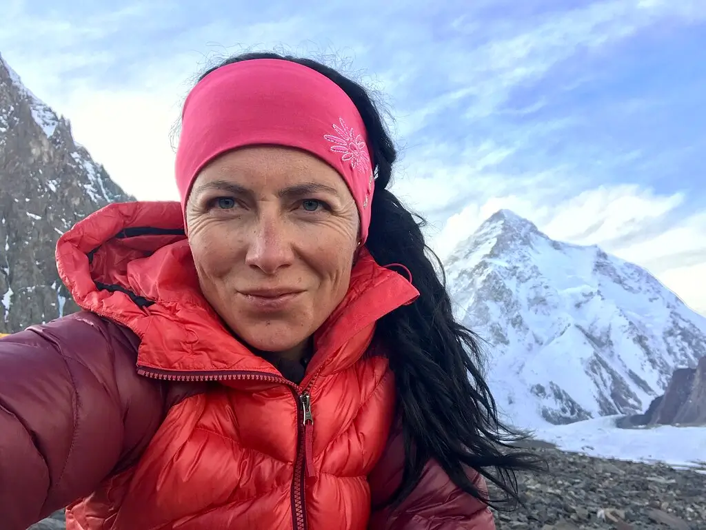 Klára Kolouchová, wearing a red down jacket and pink headband, smiles slightly while standing in a rocky alpine area with a snow-covered mountain in the background.