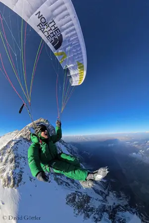 David Goettler paragliding from Nanga Parbat at 7700 meters, wearing a green down suit under a white The North Face wing, with snow-covered peaks below and clear skies above.