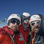 selfie of three smiling men on background of mountains, with helmets
