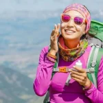 Smiling female hiker in colorful gear applying sunscreen on a sunny mountain trail to prevent sunburn at high altitude