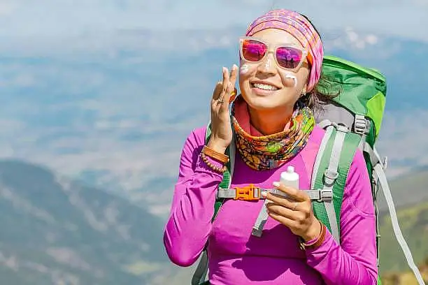 Smiling female hiker in colorful gear applying sunscreen on a sunny mountain trail to prevent sunburn at high altitude