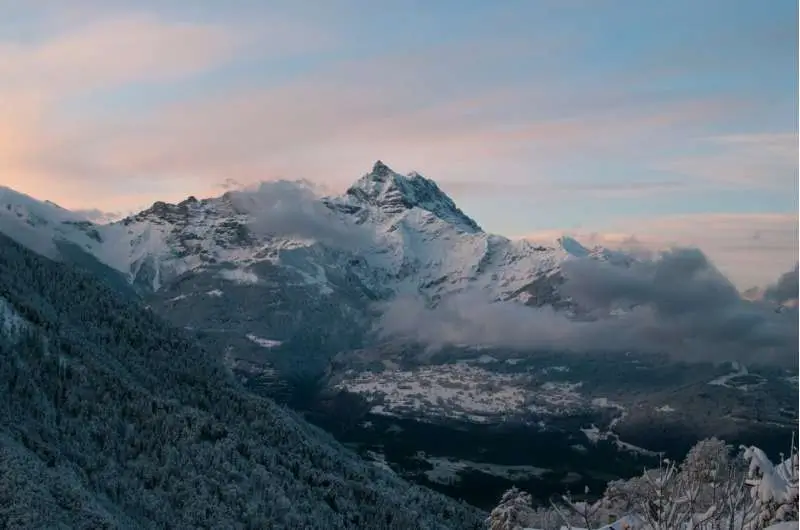 Snow-covered peaks of the Swiss Alps at sunrise, with scattered clouds drifting below the jagged summit and a forested valley visible in the foreground.