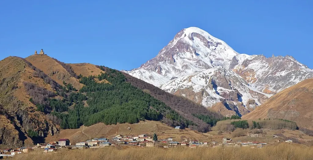 A scenic view of a small village nestled in a valley with rolling hills covered in dry grass and evergreen trees. In the background, a majestic snow-capped mountain rises sharply against a clear blue sky. A historic church with two towers sits atop a nearby hill overlooking the village.