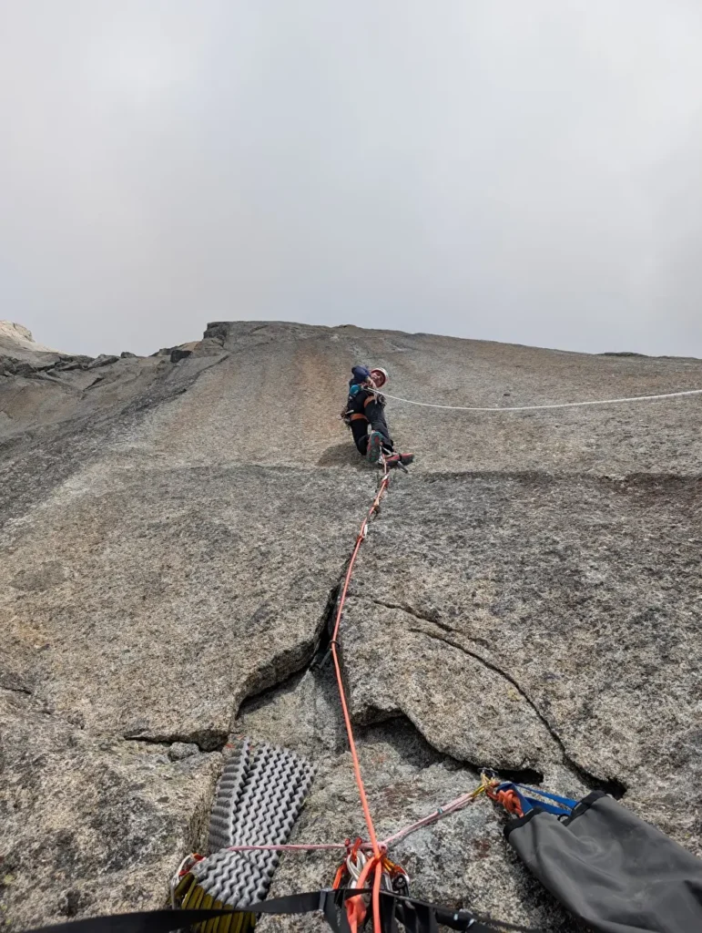 A climber in a blue helmet and dark clothing is resting on a steep granite rock face, secured by an orange rope. The climber is seated with gear, including a mat and a bag, attached to the rope. The background is a cloudy sky with the top of the rock formation faintly visible.
