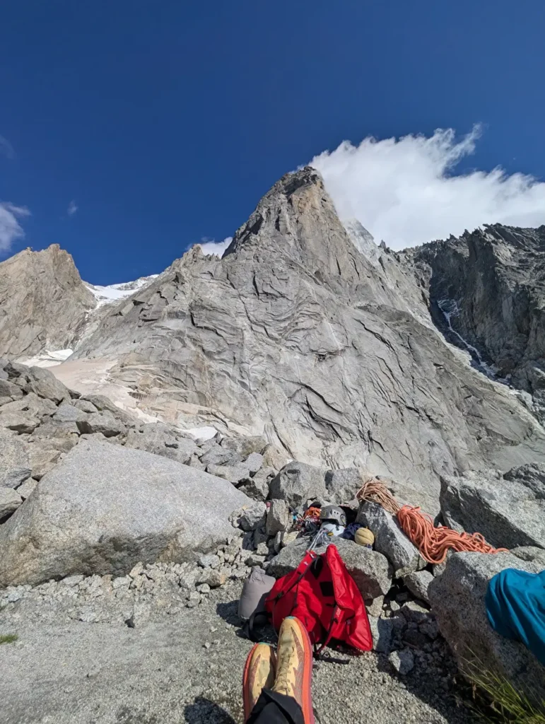 A person rests with their legs outstretched on a rocky mountainside, surrounded by climbing gear including a red jacket, a coiled orange rope, and a helmet. In the background, a steep, rugged mountain peak rises sharply against a clear blue sky with some clouds near the summit.