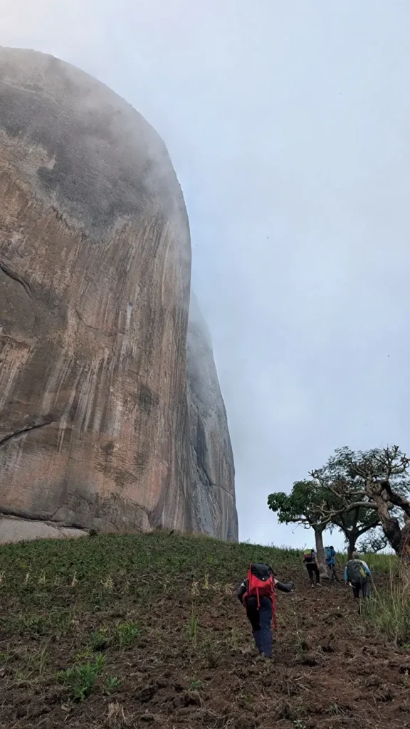 A group of hikers with backpacks ascends a steep, grassy slope toward a massive, rugged rock formation partially shrouded in mist. The scene includes a few scattered trees and a cloudy sky in the background.