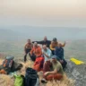 A group of hikers sits and stands on a rocky outcrop with dry grass, overlooking a misty landscape with rolling hills in the background. They are dressed in casual and outdoor clothing, some wearing jackets and hats, with backpacks and water bottles scattered around. One person gives a thumbs-up gesture.
