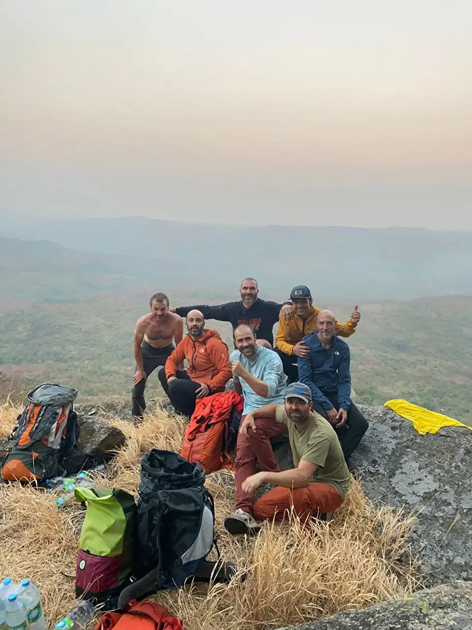 A group of hikers sits and stands on a rocky outcrop with dry grass, overlooking a misty landscape with rolling hills in the background. They are dressed in casual and outdoor clothing, some wearing jackets and hats, with backpacks and water bottles scattered around. One person gives a thumbs-up gesture.
