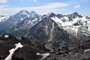 A breathtaking view of a rugged mountain range with snow-capped peaks under a clear blue sky with scattered clouds. The foreground features rocky terrain with patches of snow, while the background showcases steep, jagged mountains with visible snowfields and green valleys.