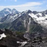 A breathtaking view of a rugged mountain range with snow-capped peaks under a clear blue sky with scattered clouds. The foreground features rocky terrain with patches of snow, while the background showcases steep, jagged mountains with visible snowfields and green valleys.