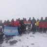 A group of people in winter gear posing together on a snowy mountain summit. They are holding several flags, including those of Argentina, India, and another with a red background and yellow symbol. The background is misty, with snow and rocks visible on the ground.