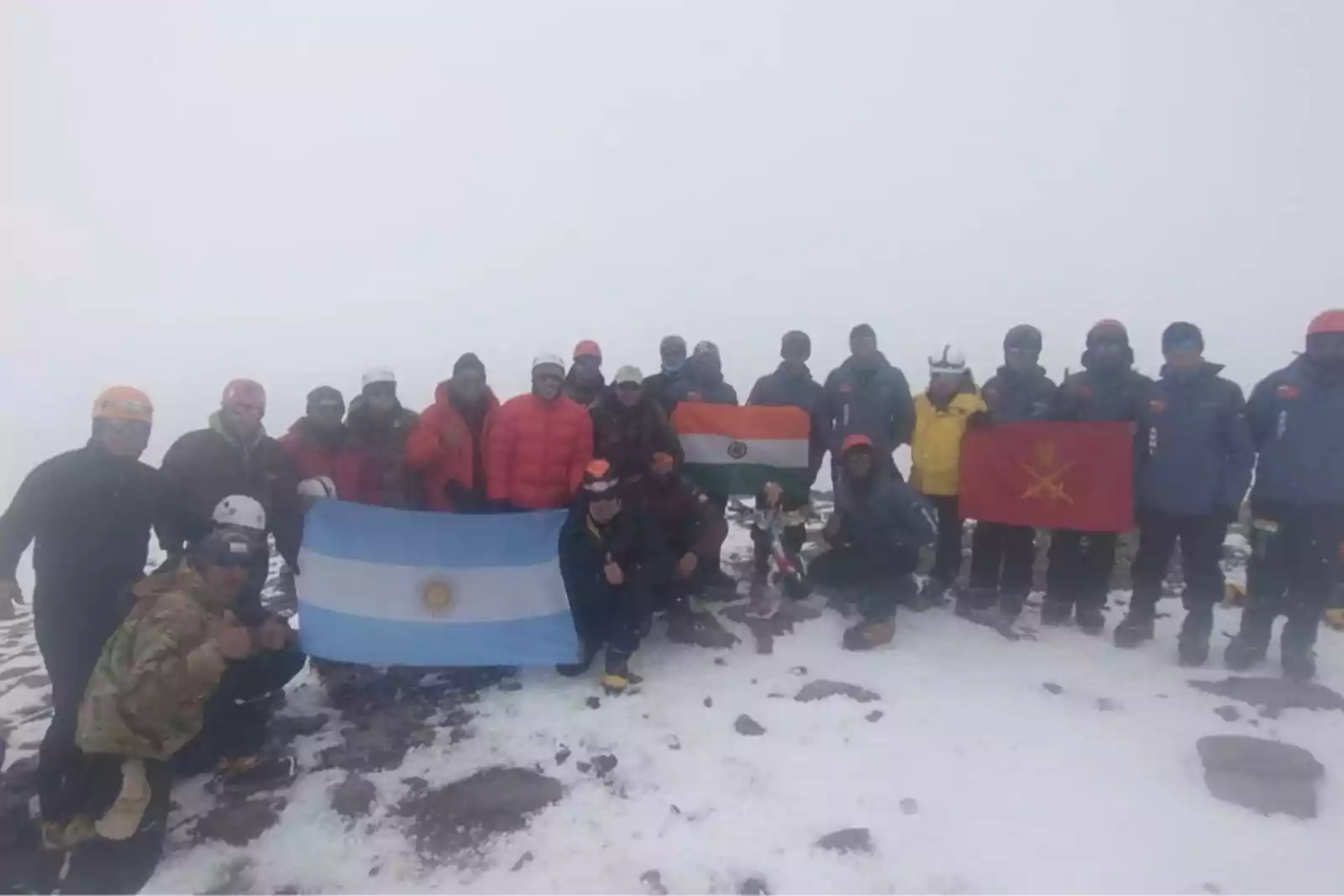 A group of people in winter gear posing together on a snowy mountain summit. They are holding several flags, including those of Argentina, India, and another with a red background and yellow symbol. The background is misty, with snow and rocks visible on the ground.