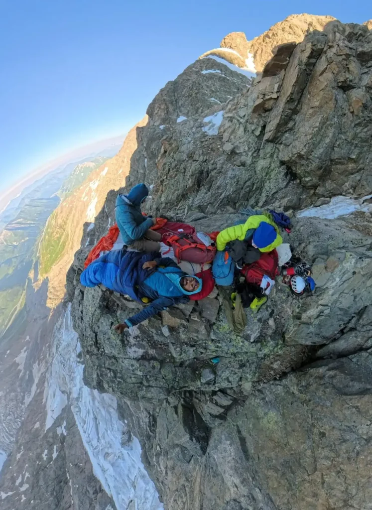 Georgian climbers bivouacking on a narrow rocky ledge high on Shkhara, secured with ropes and surrounded by steep cliffs and alpine scenery.