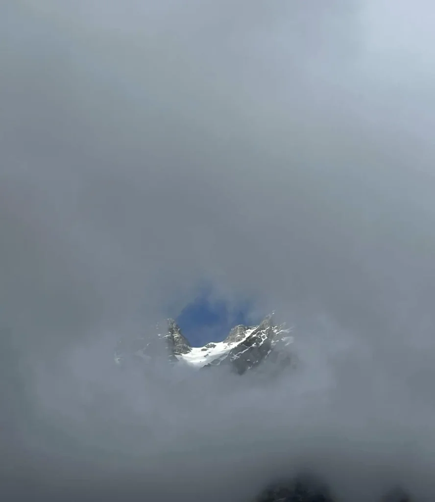 Snow-covered Ushba peaks seen through a circular break in thick clouds, revealing a patch of blue sky above the Caucasus Mountains