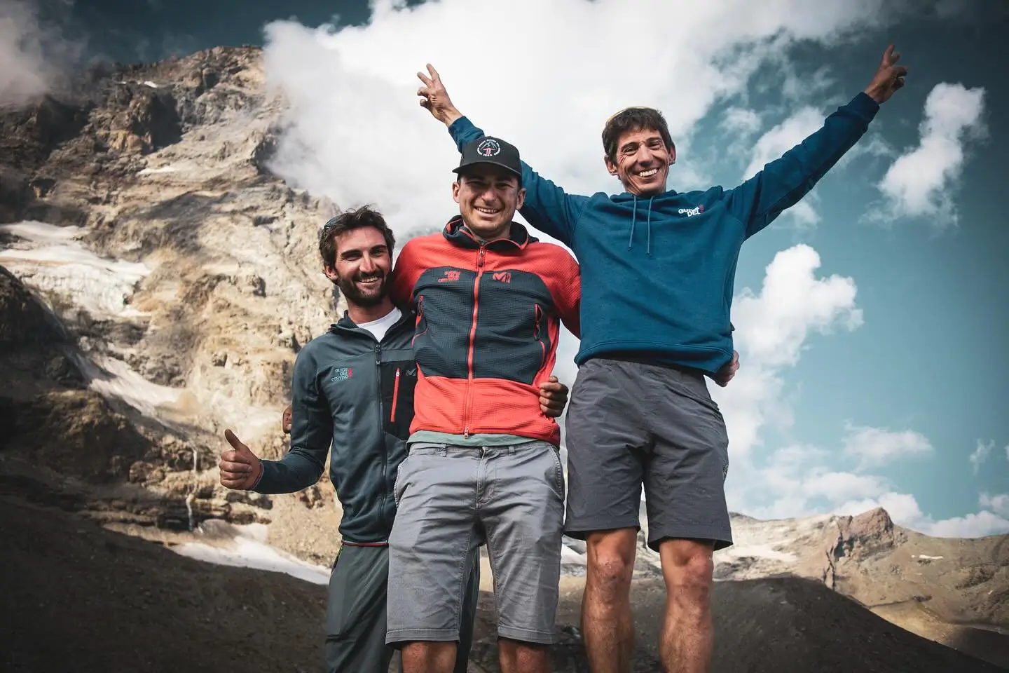 Three people standing on a rocky mountain slope with arms raised in celebration, giving thumbs-up and peace signs, with a scenic backdrop of rugged peaks, snow patches, and a partly cloudy sky.