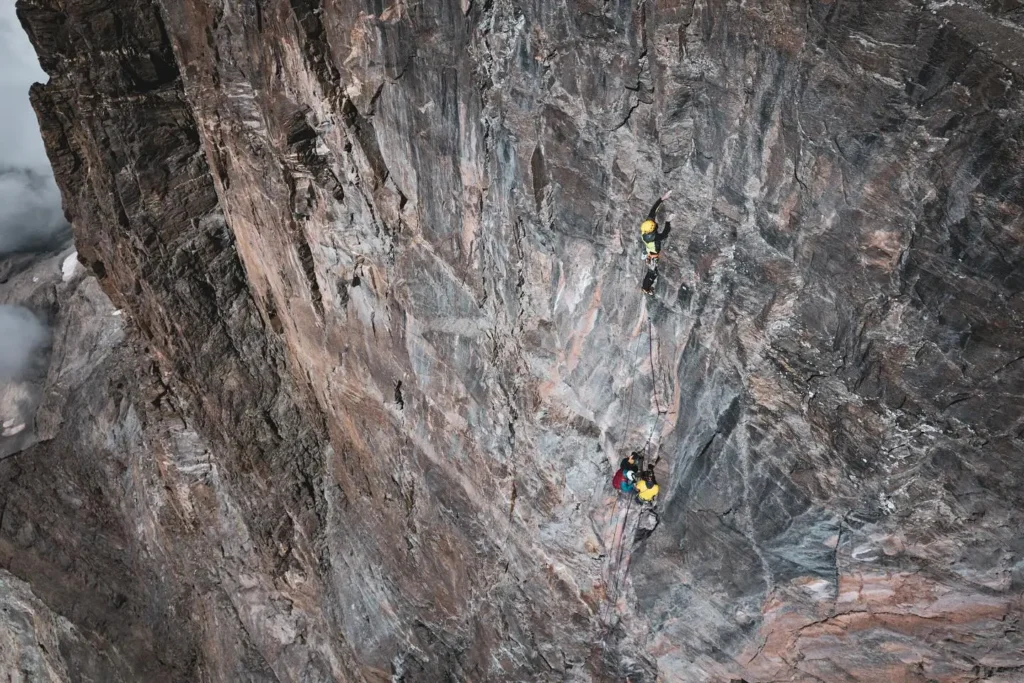 Two climbers in colorful gear and helmets ascend a steep, rugged rock face, secured with ropes, with a vast, misty mountain landscape in the background.