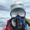 A person wearing a white climbing helmet and a red jacket takes a selfie, with an oxygen mask covering their face. The reflection in their goggles shows a stunning view of snow-covered mountains and a clear sky above the clouds.