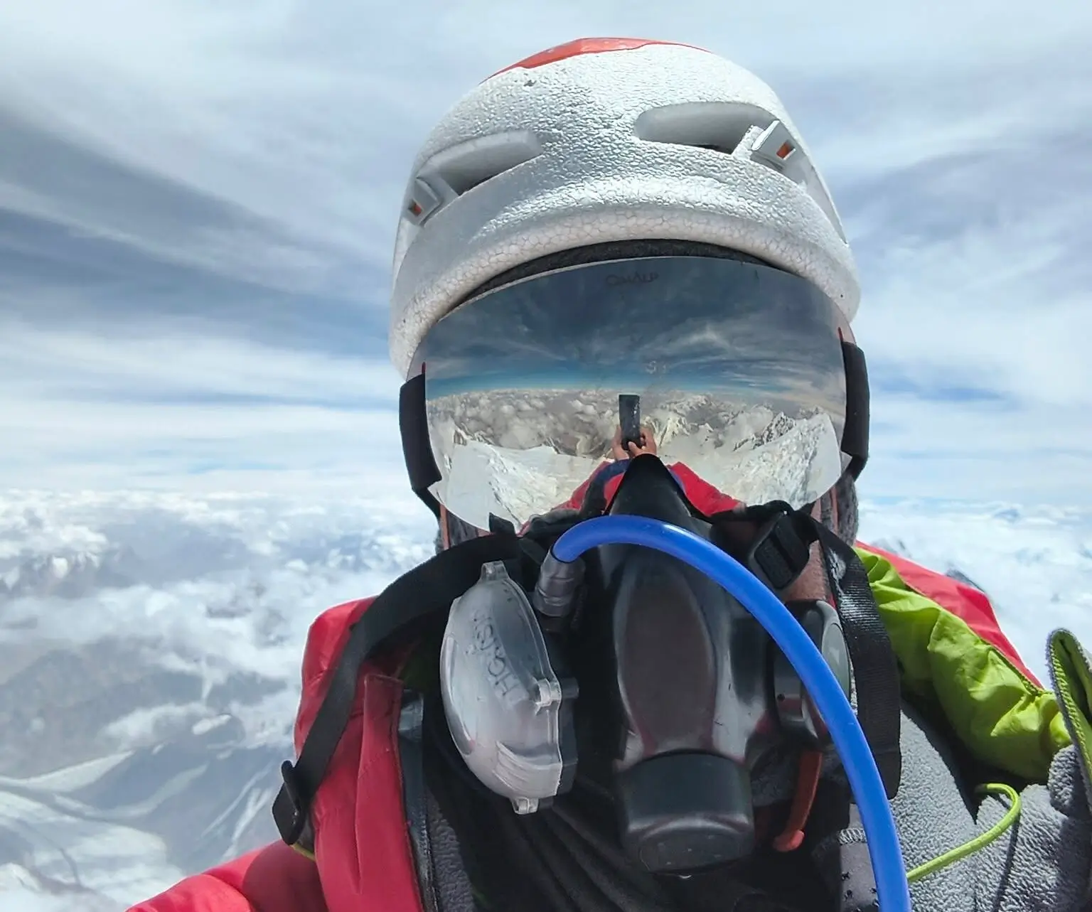 A person wearing a white climbing helmet and a red jacket takes a selfie, with an oxygen mask covering their face. The reflection in their goggles shows a stunning view of snow-covered mountains and a clear sky above the clouds.