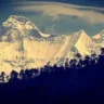 Snow‑covered twin peaks of Nanda Devi and Nanda Devi East rising above a forested ridge in Uttarakhand, India, under a dramatic sky