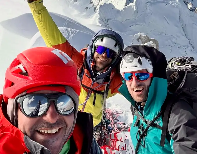 Three climbers wearing helmets and colorful jackets pose for a selfie on a snowy mountain ridge, with one raising an arm in celebration. The background features steep, snow-covered peaks under a clear blue sky.
