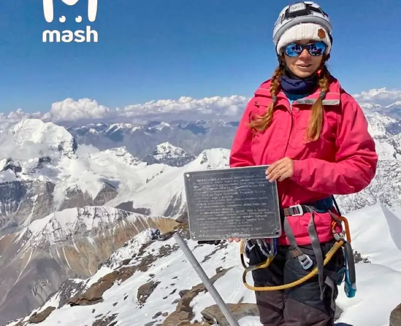 Alt text: A person wearing a bright pink jacket, climbing gear, and a white helmet with sunglasses stands on a snowy mountain peak. They hold a metal plaque with text, with a stunning backdrop of snow-covered mountains under a clear blue sky. The "mash" logo is visible in the upper left corner.