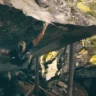 A rock climber is ascending a steep, overhanging rock face in a forested area. The climber, wearing a helmet and climbing gear, grips the rock with both hands and feet. The surrounding environment features tall trees and a rugged landscape with sunlight filtering through the canopy.