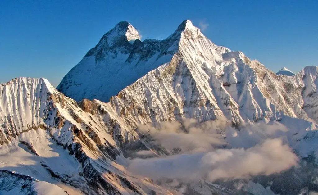 A majestic view of the snow-covered Nanda Devi mountain range under a clear blue sky, with rugged peaks and some clouds partially obscuring the lower slopes.
