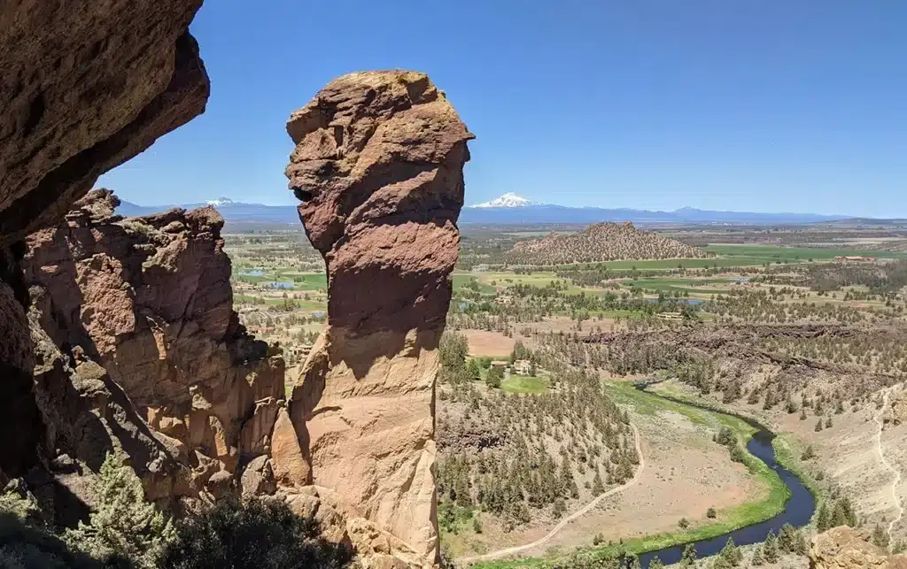 A striking rock formation rises from a cliff edge, overlooking a scenic valley with a winding river, green fields, and distant snow-capped mountains under a clear blue sky.