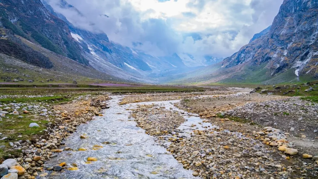 A serene valley landscape with a winding stream flowing through a rocky bed, flanked by grassy patches and surrounded by towering, misty mountains under a cloudy sky.