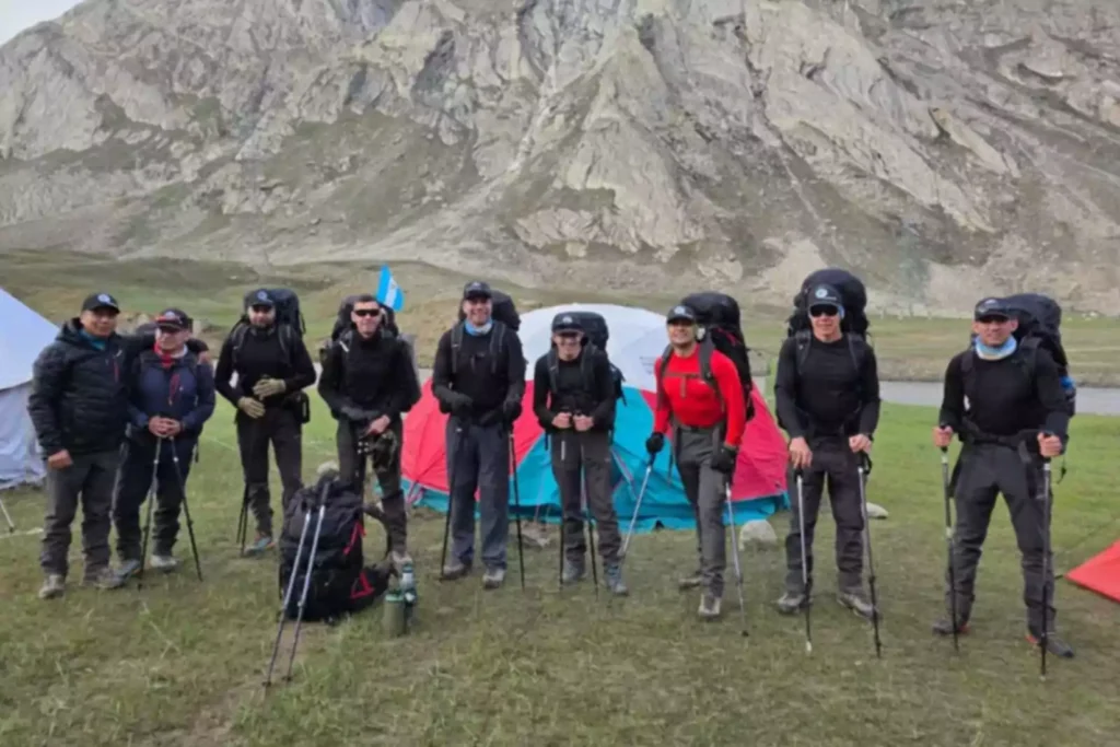 A group of hikers stands on a grassy field with rugged mountains in the background. They are dressed in outdoor gear, wearing backpacks and holding trekking poles. Some are standing near colorful tents, including a blue and red one, and a flag is visible in the background.