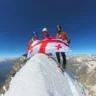 three mountaineers with helmets and sunglasses standing on summit of mountain and holding Georgia's flag. Two boys, One girl