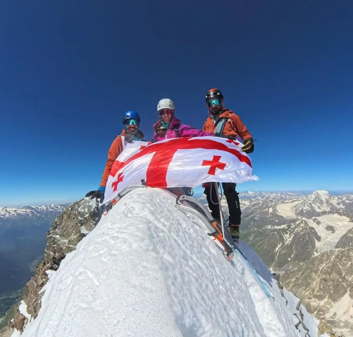 three mountaineers with helmets and sunglasses standing on summit of mountain and holding Georgia's flag. Two boys, One girl