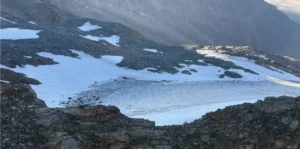 A scenic view of a snowy mountain landscape with patches of snow and rocky terrain, featuring a glacier or snowfield nestled between rugged peaks under a cloudy sky.