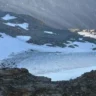 A scenic view of a snowy mountain landscape with patches of snow and rocky terrain, featuring a glacier or snowfield nestled between rugged peaks under a cloudy sky.